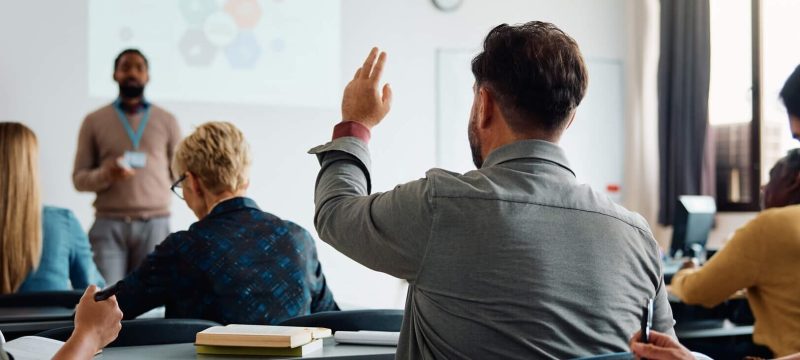 Back-view-of-student-raising-his-hand-to-answer-teachers-question-during-education-training-class-1